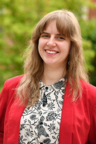 A smiling woman on a background of greenery and the red-brick Andrews House. She has her arms folded behind her and is smiling at the camera.