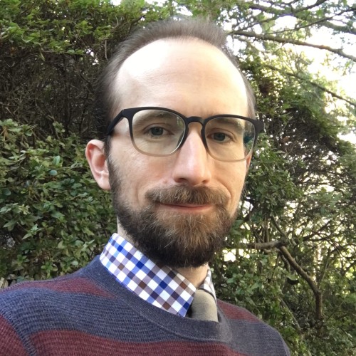 Portrait of Gregory Kimbrell, wearing a shirt of blue, brown, and white plaid and a sweater in blue and red horizontal stripes, standing before a shrub