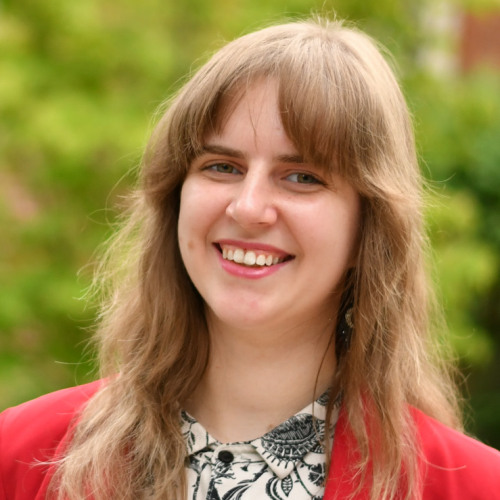 A smiling woman on a background of greenery and the red-brick Andrews House. She has her arms folded behind her and is smiling at the camera.